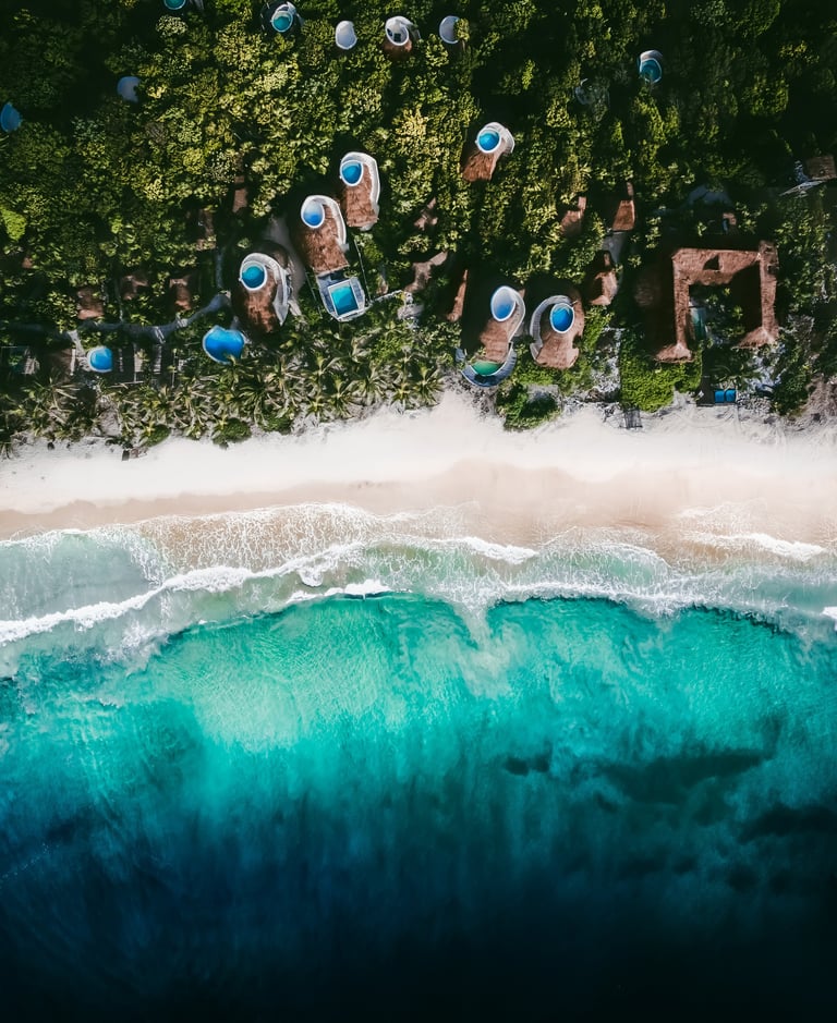 Aerial view of beach with blue umbrellas, white sand, green vegetation, and turquoise waves rolling ashore
